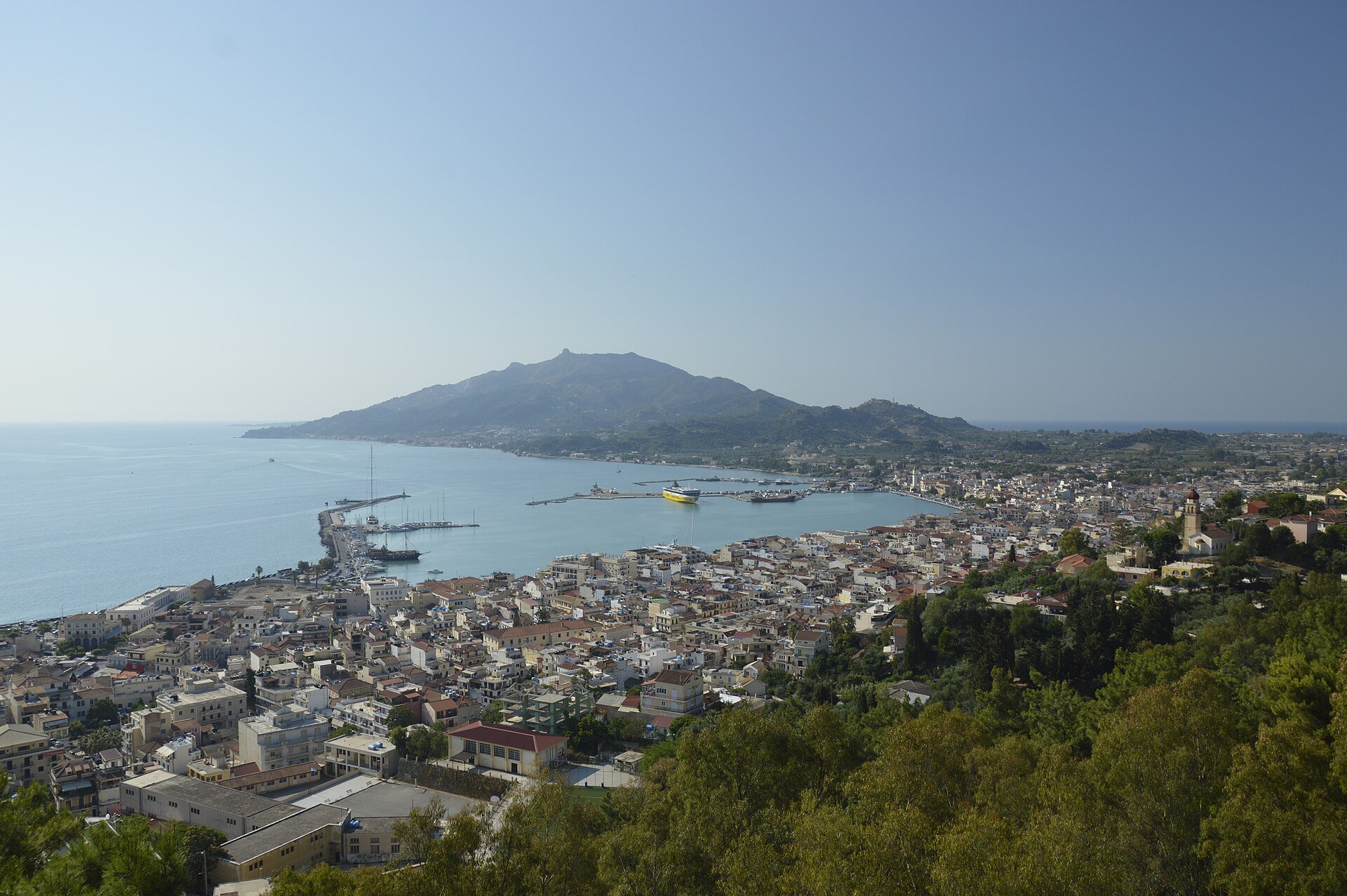 Blick von einem bewaldeten Hang über die Dächer von Zakynthos-Stadt auf Hafen und Bucht, dahinter der Berg Skopos