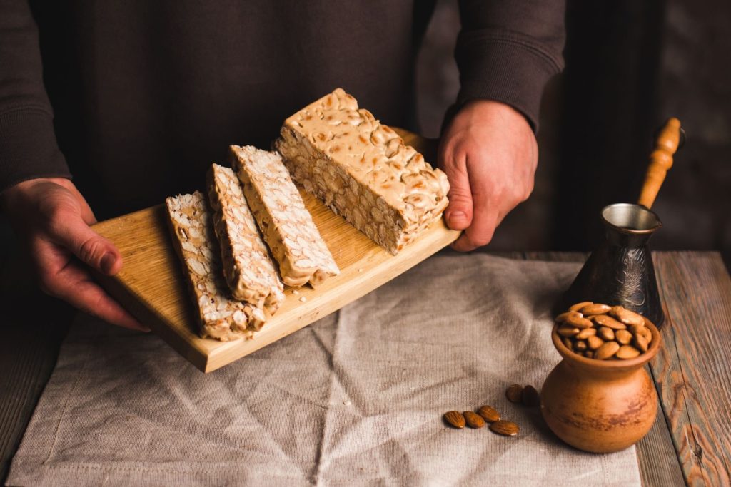 Hände halten ein Brett mit Scheiben von Mandelnougat, daneben Mandeln