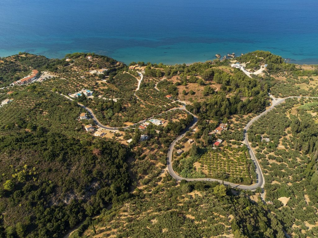 Luftbild einer kurvigen Straße durch Olivenhaine und Hügel im Nordosten von Zakynthos, dahinter das Meer