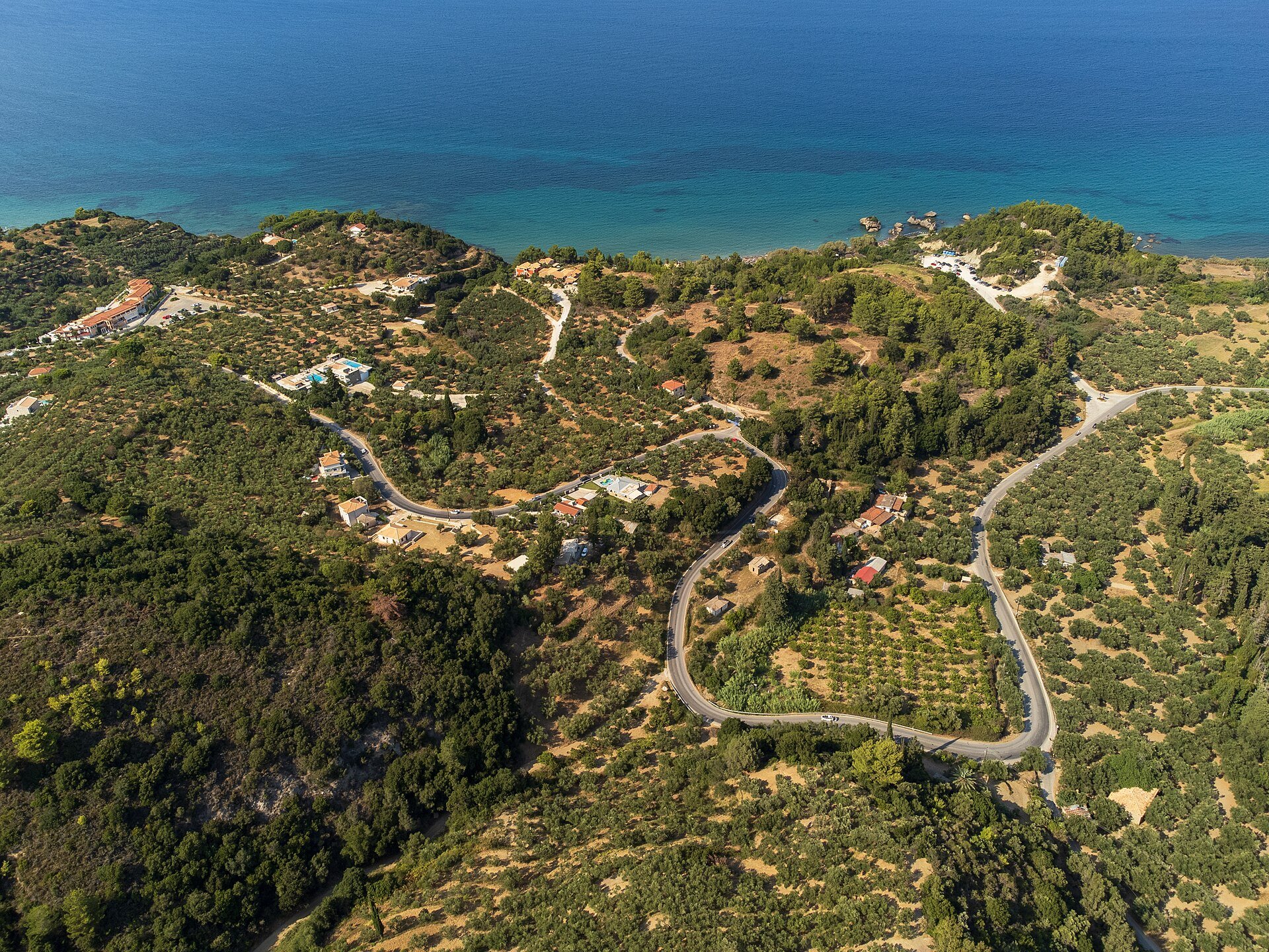 Luftbild einer kurvigen Straße durch Olivenhaine und Hügel im Nordosten von Zakynthos, dahinter das Meer