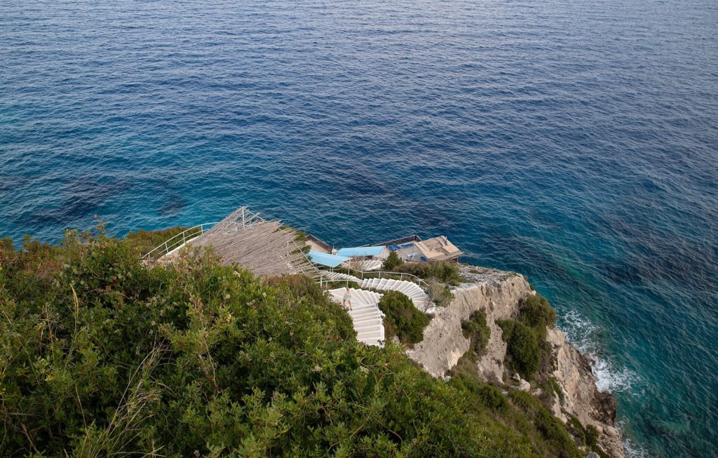 Steile Felsküste mit einer Treppe, die zwischen Gebüsch zum tiefblauen Meer hinabführt
