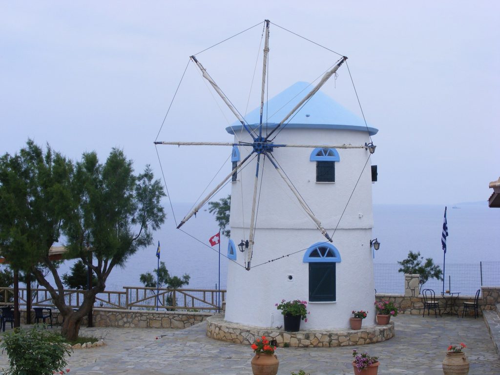 Weiße Windmühle mit blauem Dach und Flügelgerüst auf einer Terrasse über dem Meer