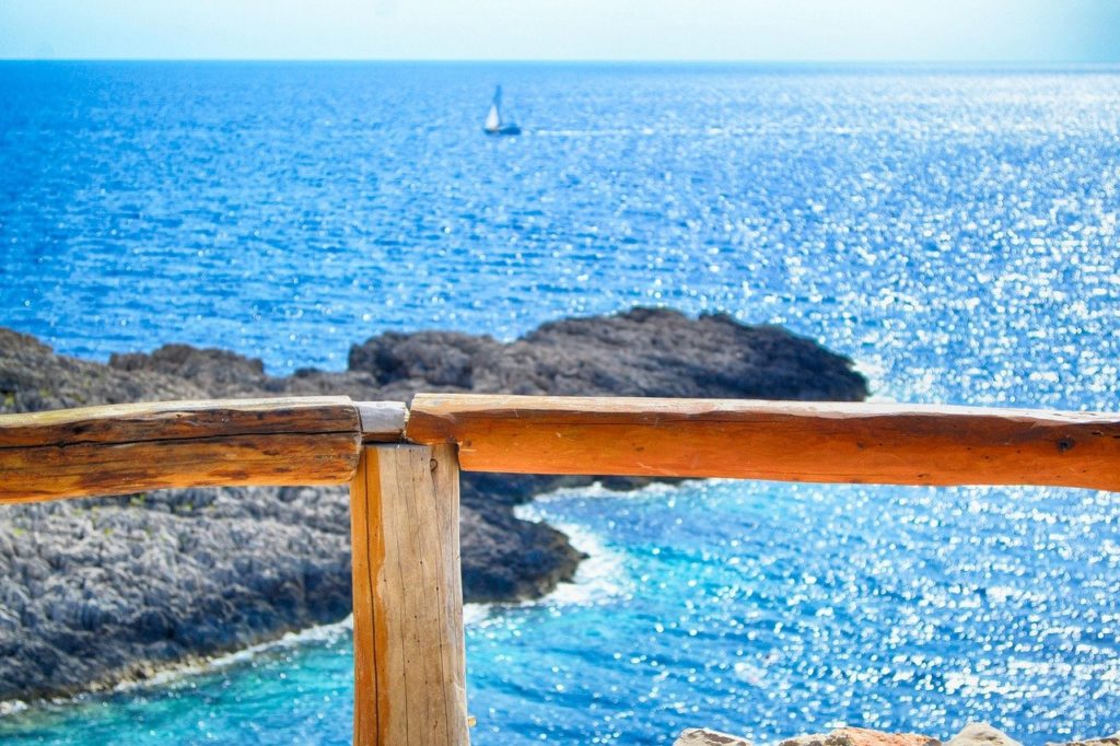 Holzgeländer vor Felsen und blauem Meer mit einem Segelboot am Horizont