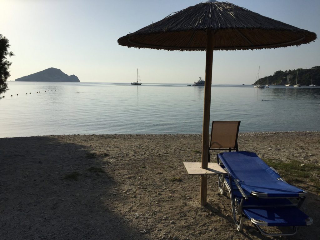 Strohschirm und Liege am Strand von Limni Keri, im Meer die Insel Marathonisi