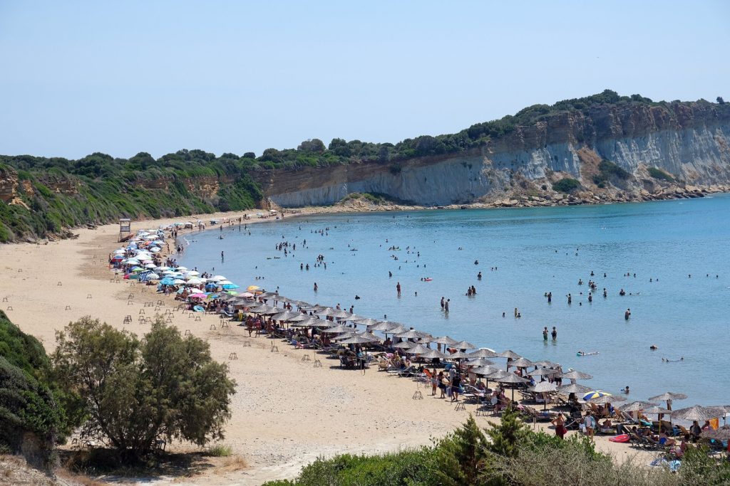 Bogenförmiger Sandstrand von Gerakas mit Sonnenschirmen, flachem Wasser und hellen Lehmklippen