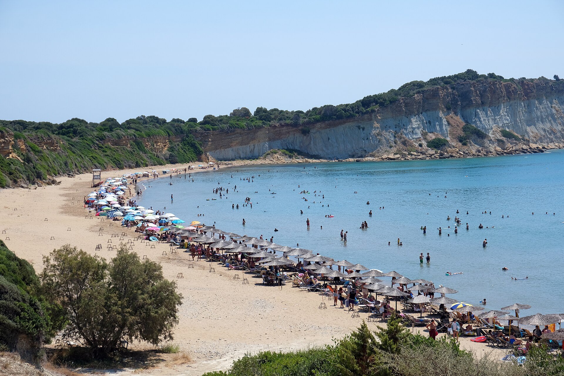 Bogenförmiger Sandstrand von Gerakas mit Sonnenschirmen, flachem Wasser und hellen Lehmklippen