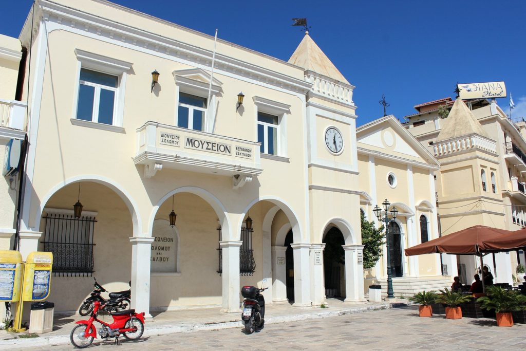 Helles Gebäude des Solomos-Museums mit Arkaden am Markusplatz, rechts die Kirche San Marco
