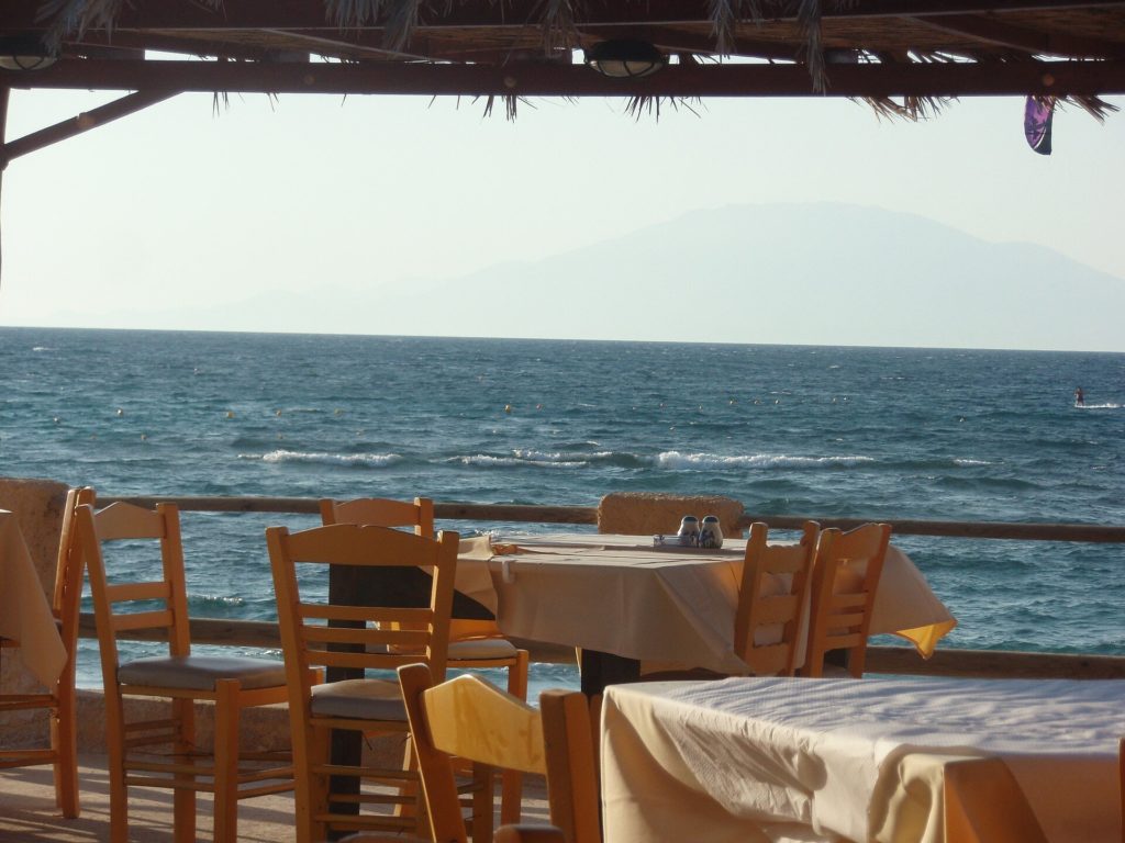 Blick von einer schattigen Terrasse mit Holzstühlen am Strand von Alykes auf das bewegte Meer