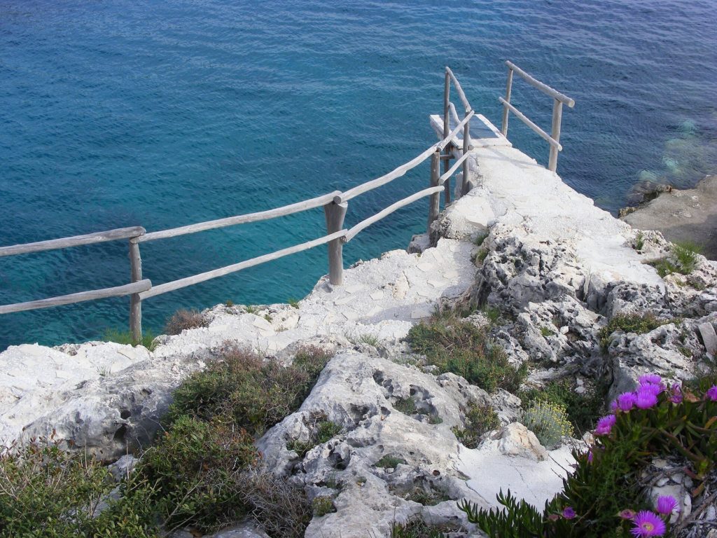 Felsige Steilküste mit Holzgeländer und Stufen über dem türkisblauen Meer in Porto Roxa