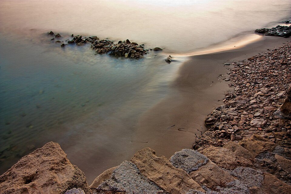 Sandstrand mit Felsen im flachen Wasser bei Amoudi im Abendlicht